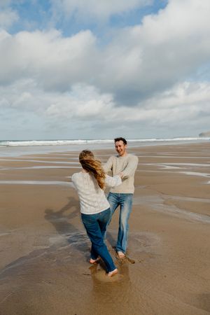 Couple dancing barefoot on a sandy beach with ocean waves and cloudy sky in the background.