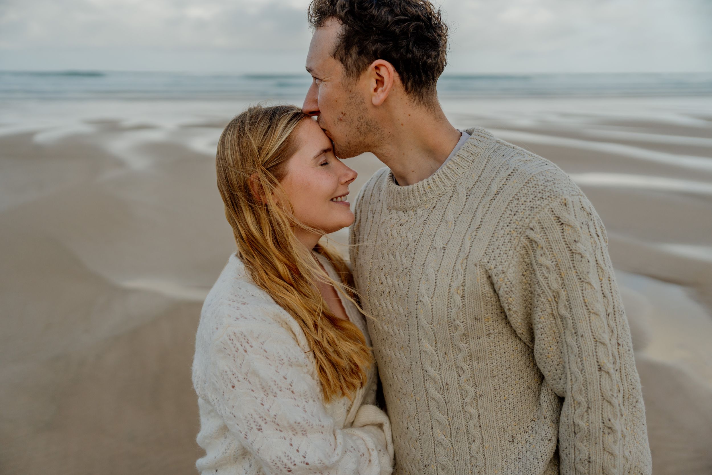 A couple in cozy cable-knit sweaters share a tender moment on a sandy beach with ocean waves in the background.