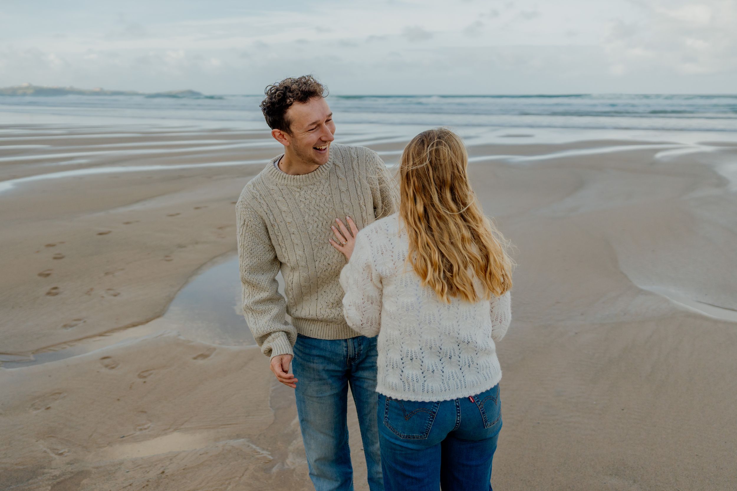 Couple laughing together on a sandy beach, wearing cozy knit sweaters and jeans on a cloudy day.