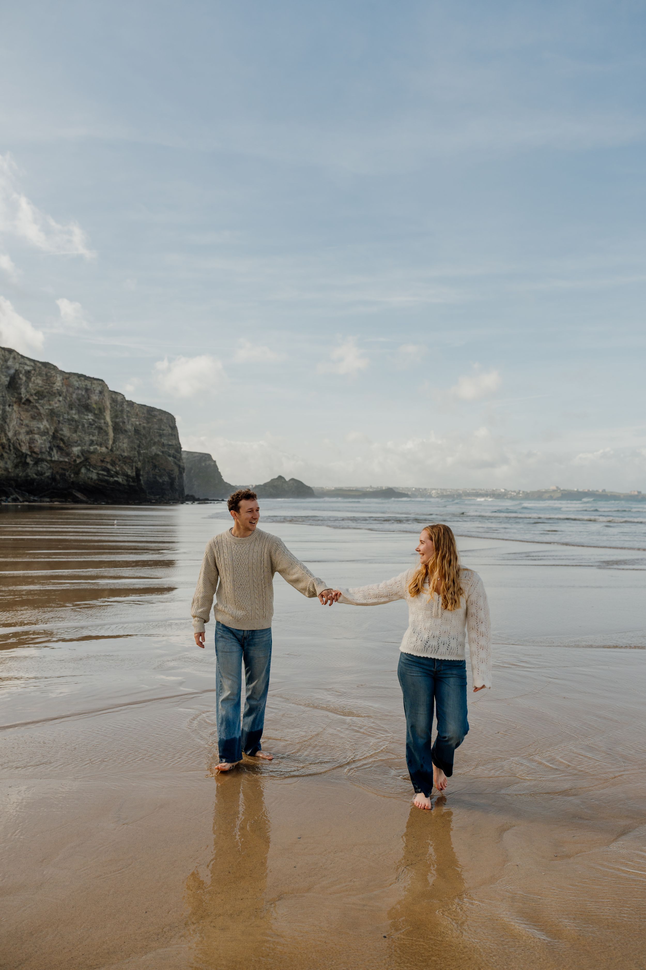 Couple holding hands walking barefoot on a sandy beach with ocean waves and dramatic cliffs in the background.