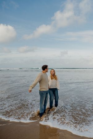 Couple in matching cream sweaters and jeans walking hand in hand along a sandy beach with ocean waves.