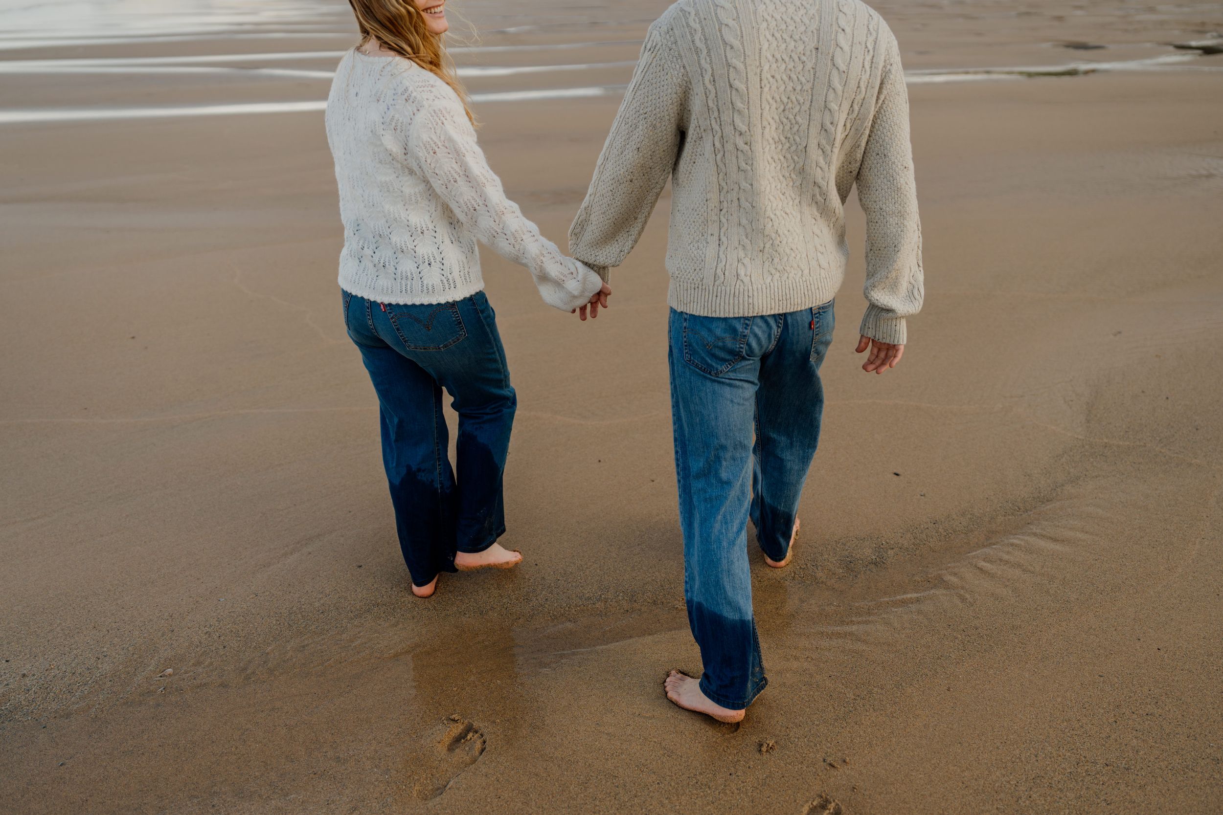 Couple holding hands walking barefoot on a sandy beach wearing white knit sweaters and blue jeans.