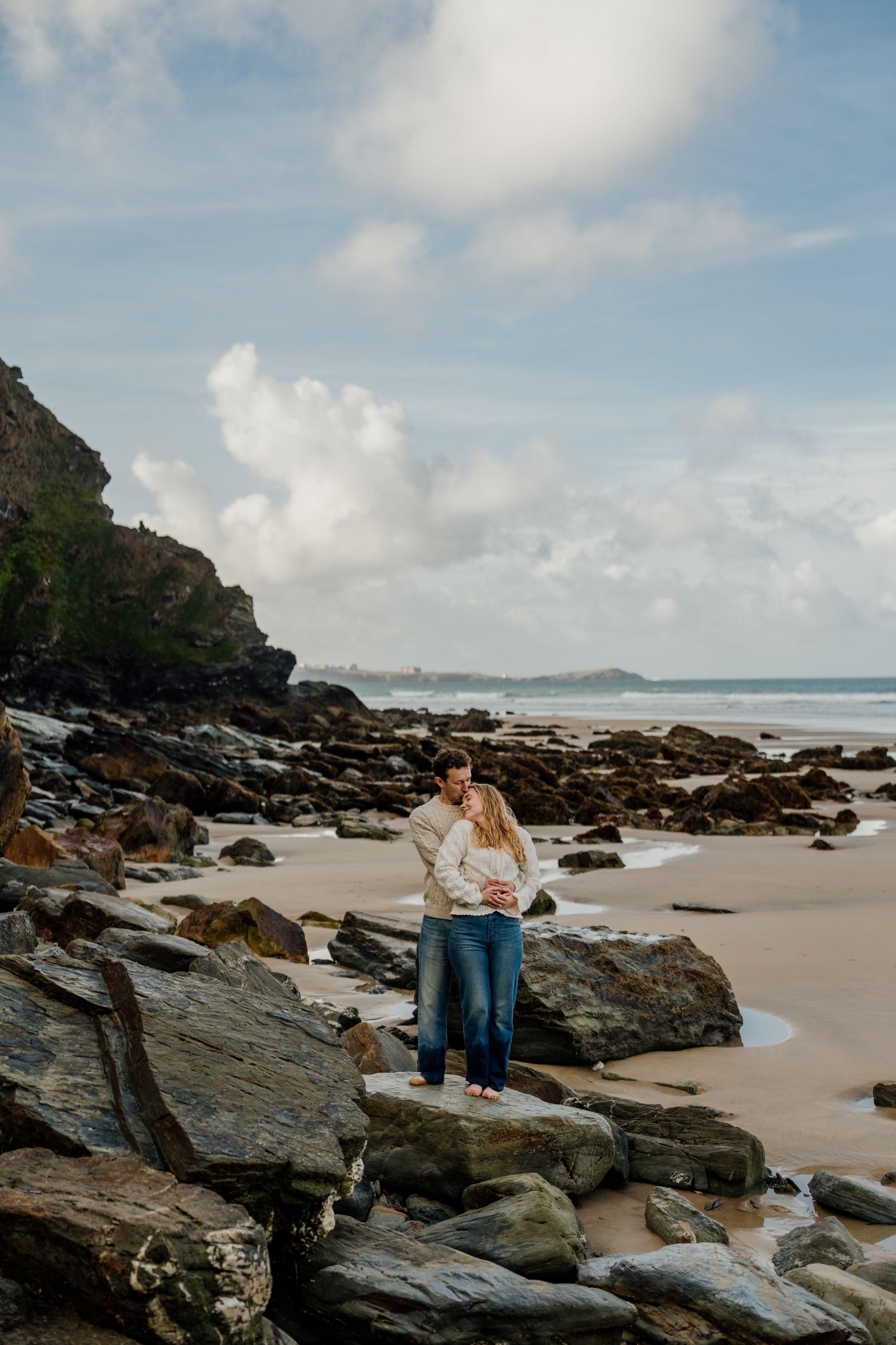 Couple embracing on rocky beach with sandy shore and cloudy sky backdrop.