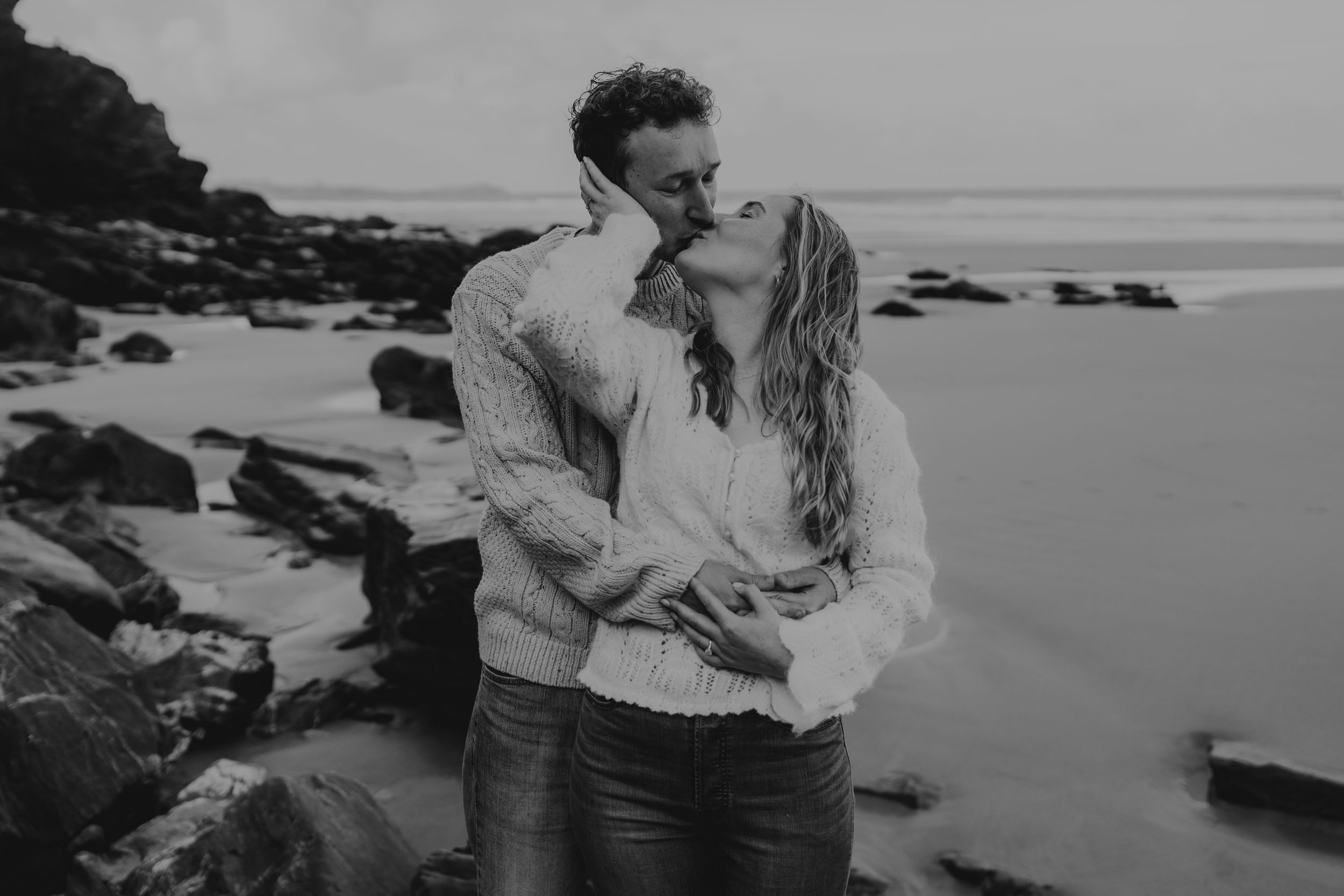 Black and white photo of a couple sharing a romantic kiss on a rocky beach with ocean waves in the background.