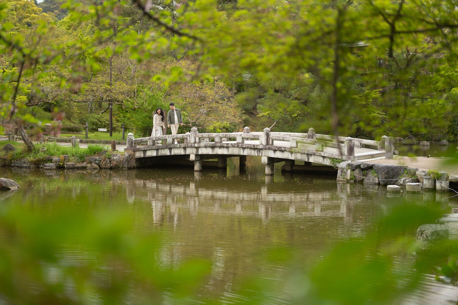 Couple in wedding attire standing on a wooden arched bridge over a tranquil pond in a lush green garden.