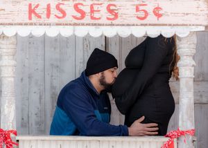 Couple sharing a kiss at a rustic 'Kisses 5 cents' booth, with the woman's pregnant belly visible.