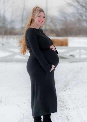 Pregnant woman in black long-sleeve dress cradling baby bump outdoors in snowy winter landscape.