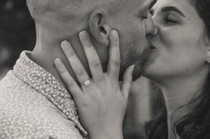Black and white photo of a couple kissing, woman showing off diamond engagement ring on man's neck.
