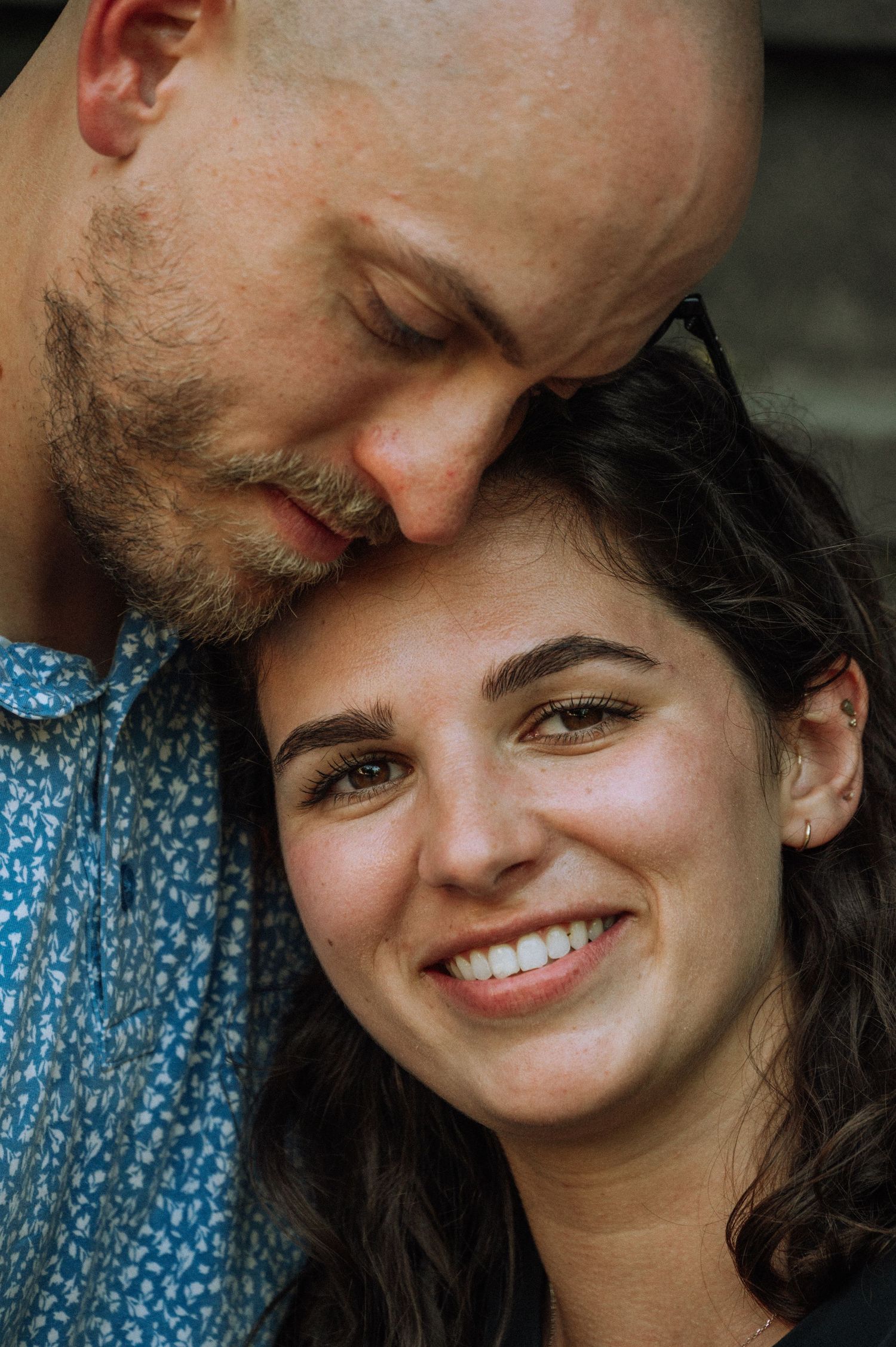 A bald man with stubble tenderly rests his head against a smiling young woman with curly brown hair outdoors.