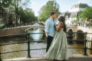 Couple sharing a romantic moment on a canal bridge in Amsterdam, with historic buildings and waterways in the background.