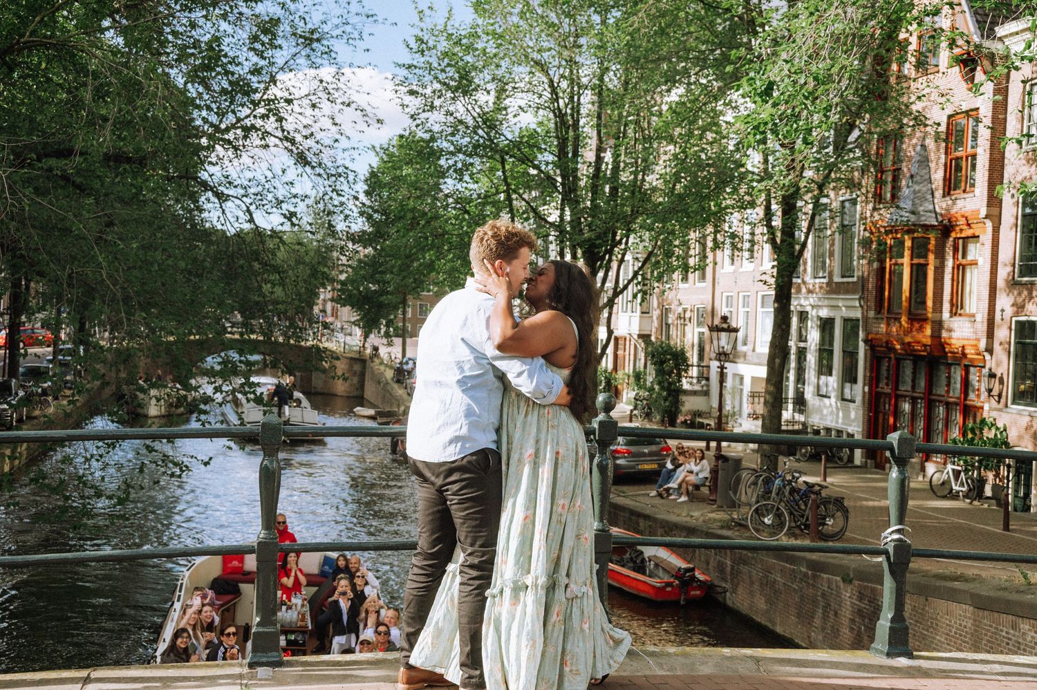 Couple sharing a romantic kiss on a bridge over an Amsterdam canal surrounded by lush trees and colorful boats.