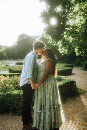 Couple kissing in a sunlit garden park, woman in floral mint dress holding hands with man in light blue shirt.