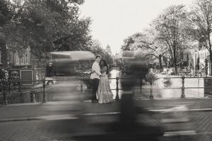 Black and white photo of a couple embracing on a bridge in Amsterdam with motion-blurred cyclists passing by.