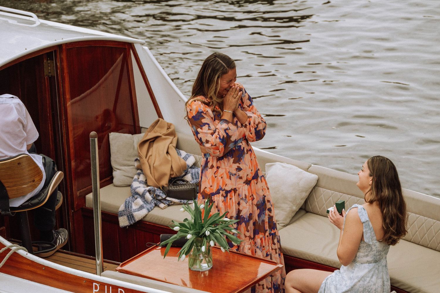 Two women on a luxury boat on the water, one in a floral dress and another in a white dress holding a drink.