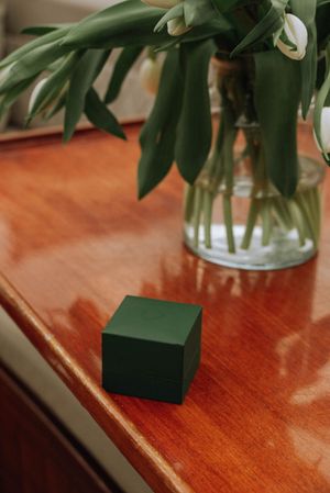 A small green jewelry box sits on a polished wooden surface beside a glass vase with white tulips.