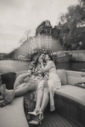 Black and white photo of two women laughing and embracing on a boat along a scenic canal in Amsterdam.