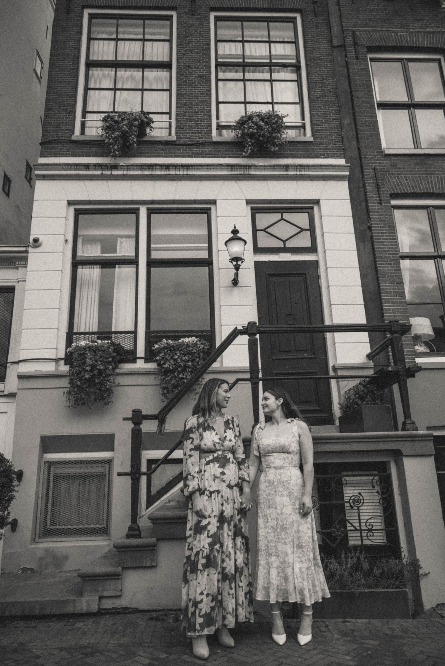 Two women in floral dresses stand together outside a classic Amsterdam canal house in black and white photo.