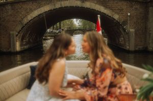 Two women sit on a boat gliding under a brick arch bridge on an Amsterdam canal.