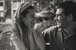 Black and white photo of a smiling couple sharing a joyful moment outdoors, woman covering her face with hands.