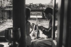 Black and white photo of a couple on a boat enjoying drinks on a canal with a bridge in the background.