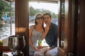 A smiling couple poses on a boat, woman showing engagement ring, with drinks and waterway visible through window.