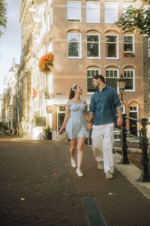 A couple holds hands walking along a sunny Amsterdam canal street, woman in blue dress, man in white pants.