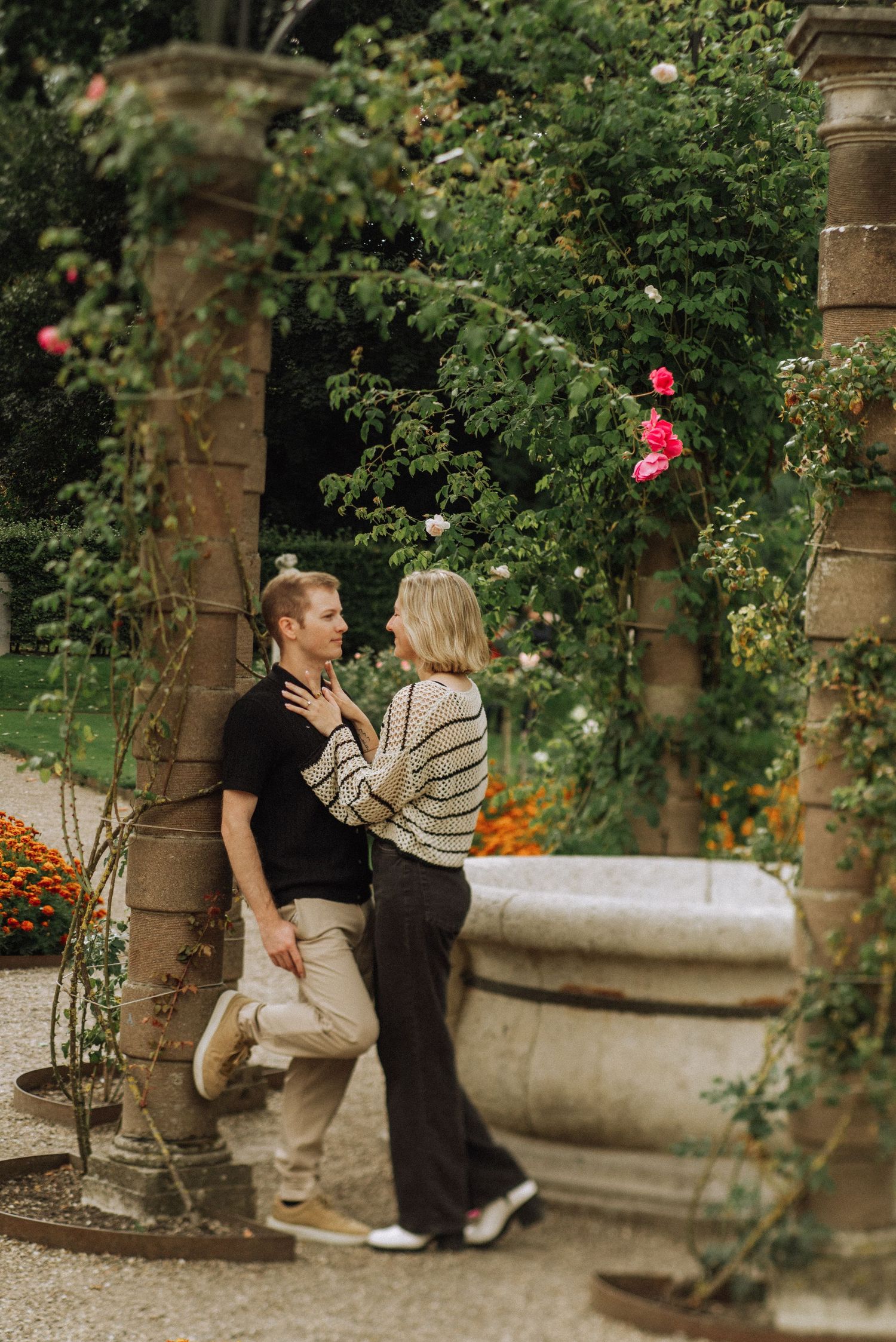 Couple sharing a romantic moment in a lush garden with climbing roses and stone columns.