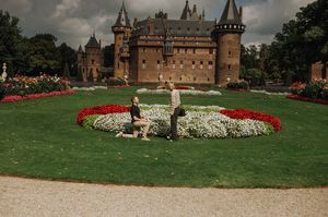 Couple sitting in manicured gardens with red and white flowers before a grand medieval brick castle under cloudy skies. Kasteel de Haar