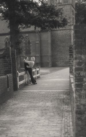Black and white photo of a couple embracing on a cobblestone path beside a historic brick church.