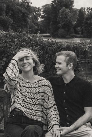 A smiling couple sits outdoors on a bench surrounded by lush greenery in a candid black and white portrait.