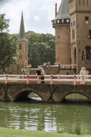 Couple embracing on a stone bridge over a moat beside a medieval castle with towers and a church spire.