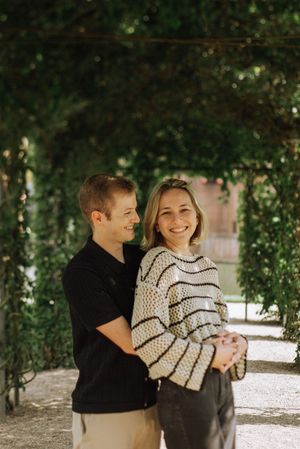 A smiling couple embraces under a lush green garden archway, the woman in a striped sweater looking at the camera. Kasteel de Haar