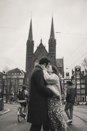 Black and white photo of a couple kissing in Amsterdam with a Gothic church and city street in the background.