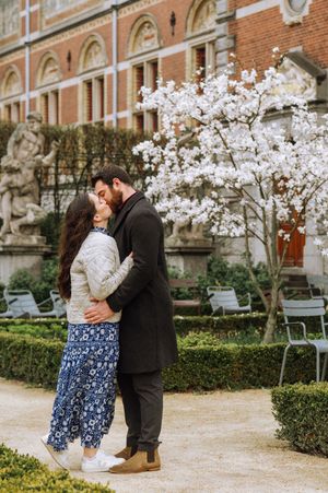 Couple embracing and kissing in a garden with blooming white magnolia tree and historic brick building backdrop.