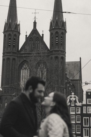Couple sharing a romantic kiss in black and white with Gothic church towers in Amsterdam background.