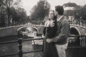 Black and white photo of a couple embracing on a bridge over an Amsterdam canal with boats and trees in background.