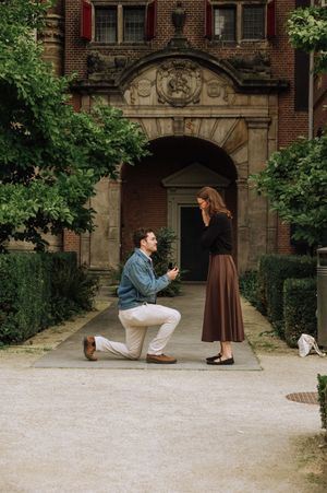 Man proposes on one knee to woman in brown skirt outside ornate brick building entrance surrounded by green trees.