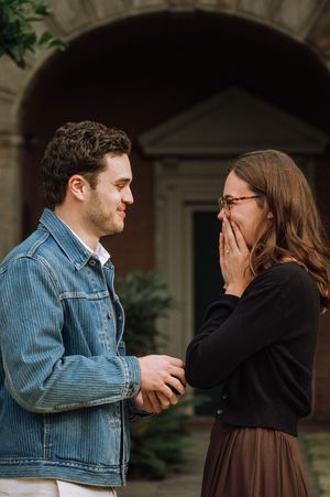 A man in a denim jacket proposes to a surprised woman in glasses who covers her mouth outdoors.