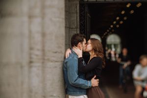 A couple shares a romantic kiss against a stone column in an elegant archway with bokeh lights in the background.