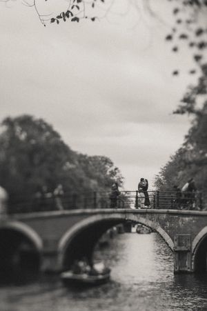 Black and white photo of a couple kissing on a bridge over a canal, surrounded by trees and a cloudy sky.