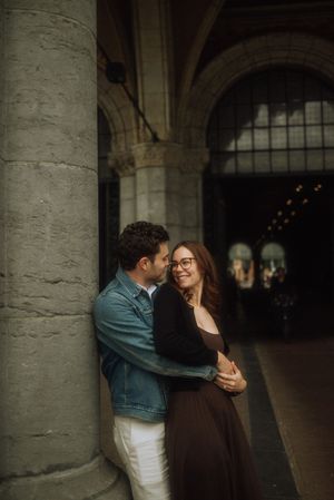 Couple embracing romantically by a stone column in an elegant arched building, woman in brown dress smiling warmly.