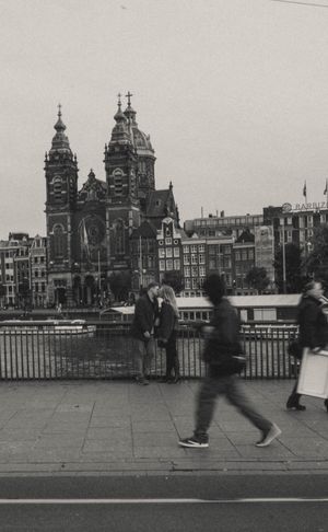 Black and white photo of pedestrians walking near a canal in Amsterdam with the Basilica of Saint Nicholas in the background.
