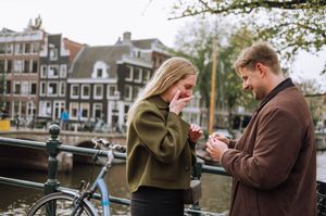 Couple sharing a romantic marriage proposal moment on an Amsterdam canal bridge with bicycles and historic buildings.