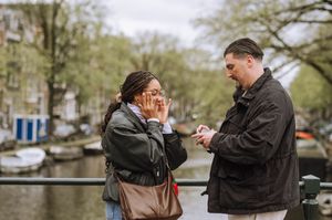 Man proposing to surprised woman on Amsterdam canal bridge, her hands covering her face in joy.