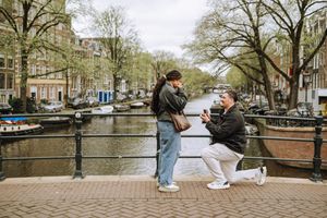 Man proposing to woman on a bridge over Amsterdam canal with boats and trees in background.