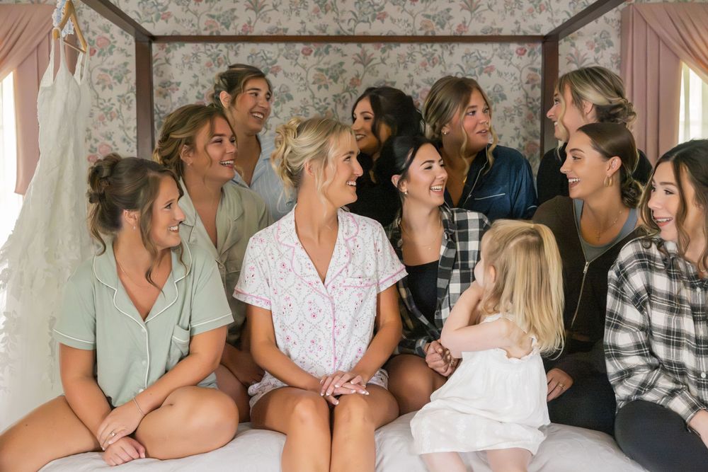 Bride in white pajamas laughing with bridesmaids and friends on a bed during wedding morning preparations.