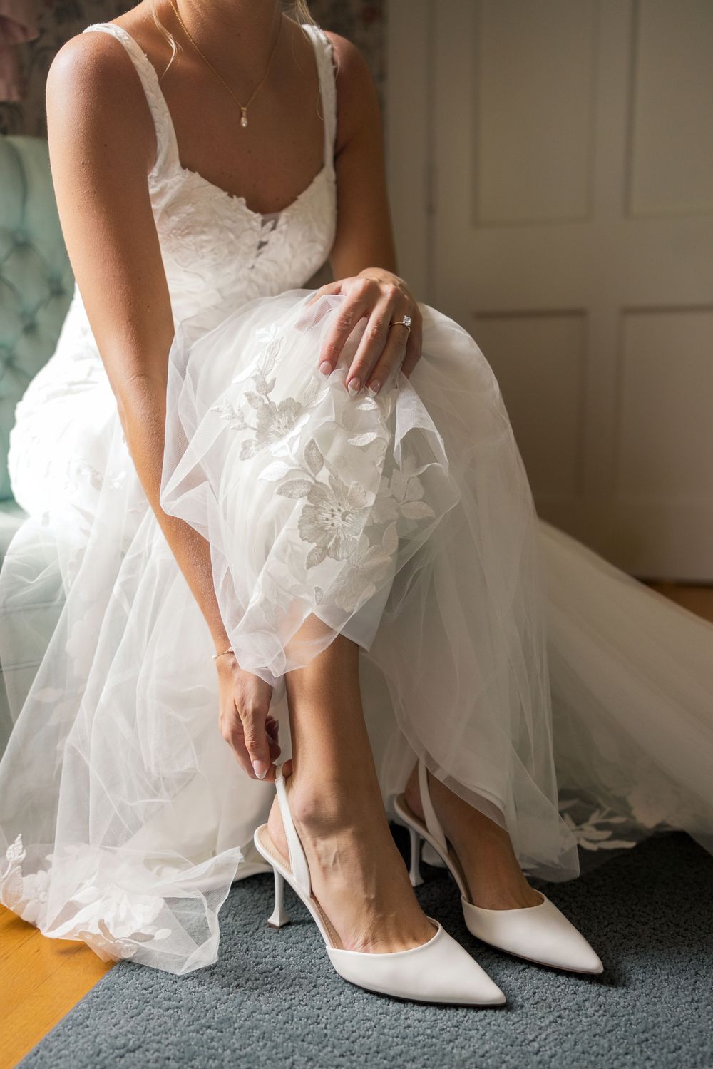 Bride in white lace wedding gown sitting and putting on elegant white satin pointed-toe heels.