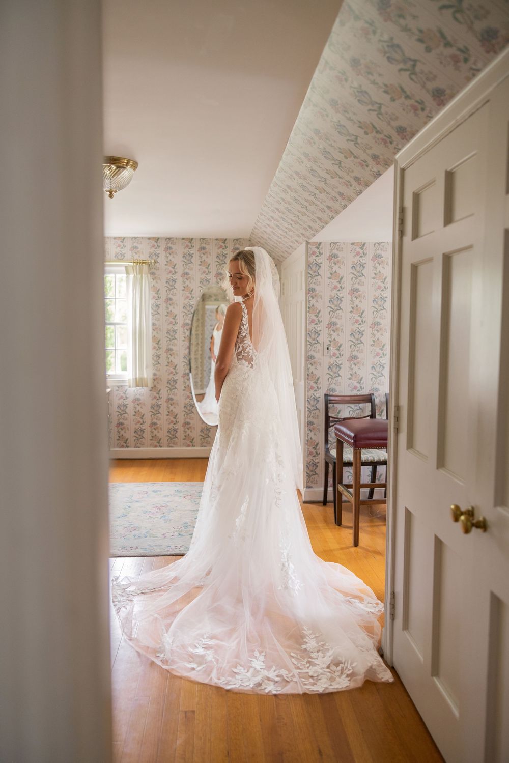 Bride in elegant lace wedding gown with long train and veil stands in a floral wallpapered room on hardwood floors.