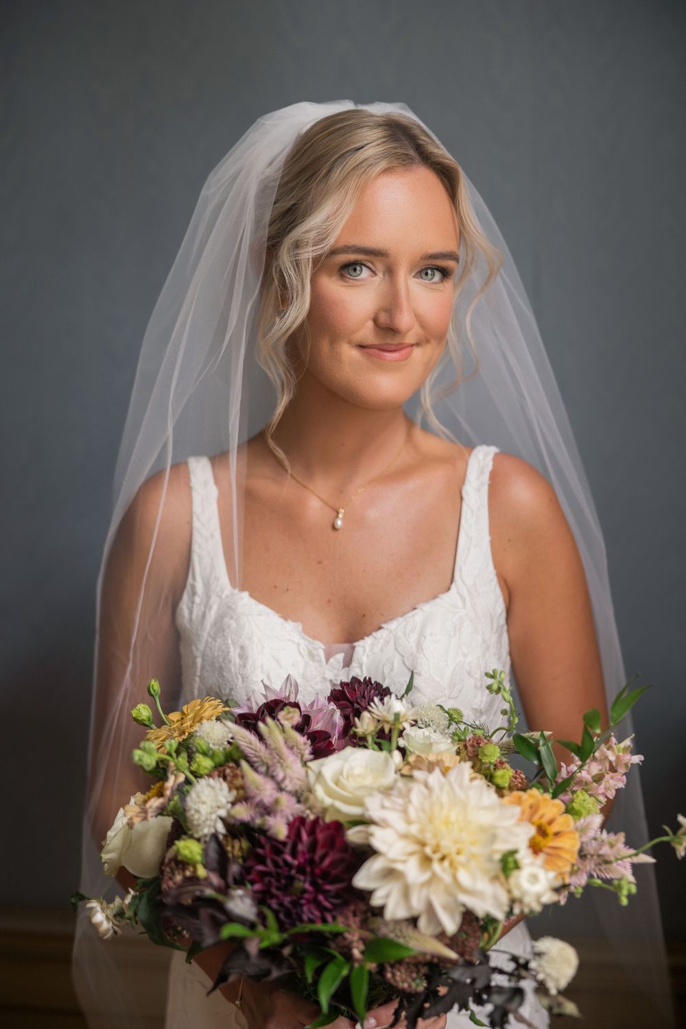 Bride in white lace dress and veil holds colorful bouquet with dahlias and wildflowers, posing for portrait.