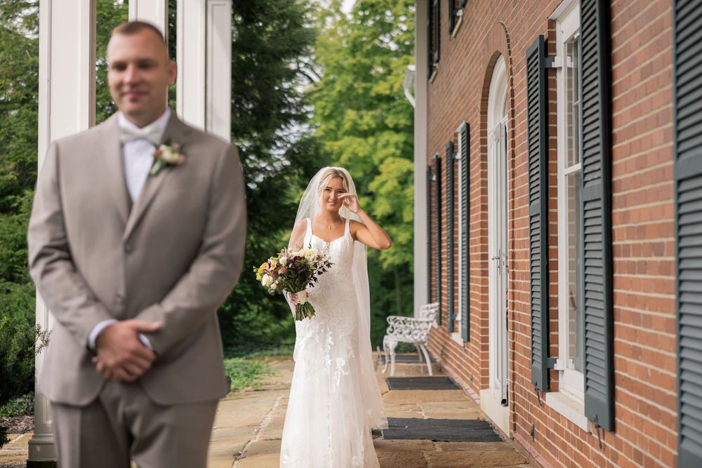 Emotional bride in white gown crying during first look with groom in tan suit outside brick building.